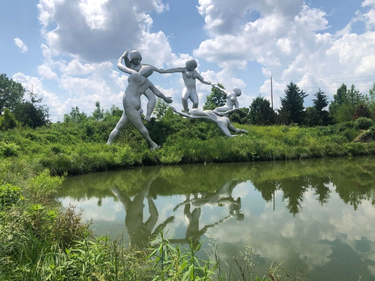 Large metal statue of five women holding hands and dancing in front of a pond.