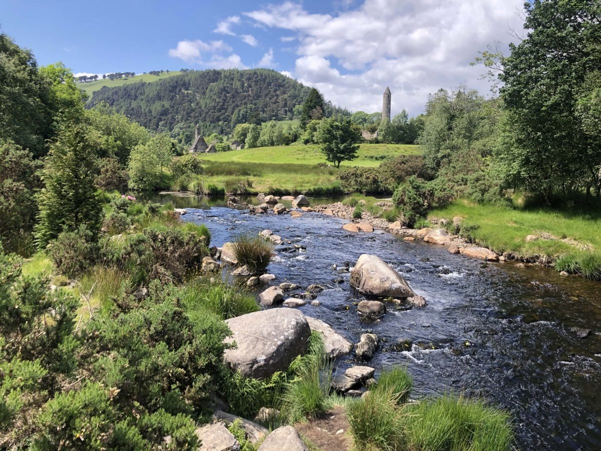 A wooded area with a river, stones, and a tall stone tower in the background.