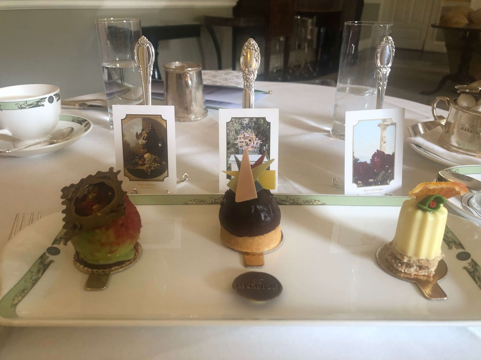 Three fancy pastries on a rectangular plate with three small pictures behind them, on a tea table.